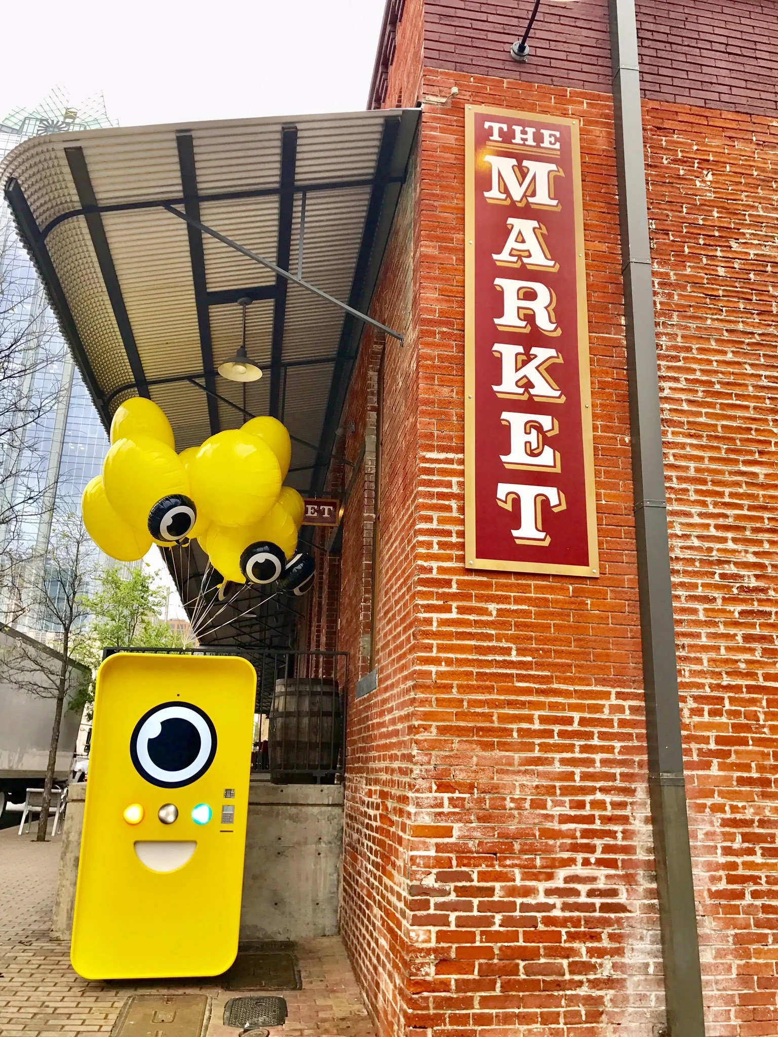 Yellow Snapbot vending machine with balloons beside a brick wall bearing a vertical red THE MARKET sign